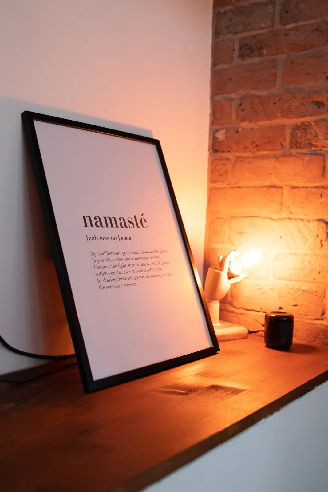 Dettaglio d'arredo su una mensola in legno: una stampa incorniciata con la parola Namasté, illuminata da una lampada, contro il muro di mattoni.